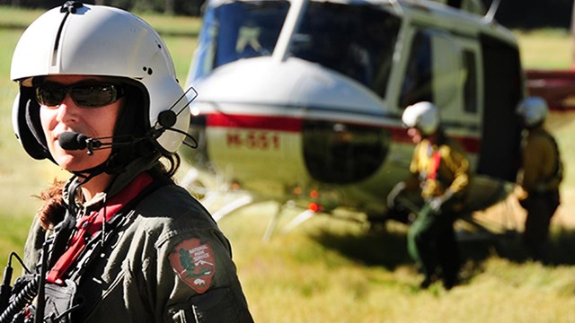 A helicopter crew member stands in the foreground with two other people and a helicopter in the back